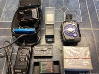 Photo showing assorted vintage handheld electronic devices and accessories including Sony digital voice recorder, Panasonic battery charger, cassette adapters and others, laid out on a tiled floor.