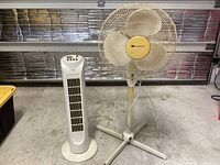 Two used floor fans, one white tower fan and one white pedestal fan, placed on concrete floor in garage or basement setting.