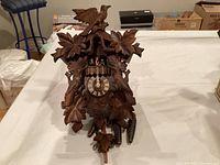 Front view of the German cuckoo clock showing carved wooden leaves, bird atop, clock face with Roman numerals, two figurines, and three pinecone-shaped weights.