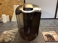 Black Keurig K50 coffee maker with water reservoir, accessory parts in sealed bag, and user manual booklet on tiled countertop.