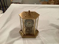 Full view of the gold tone hexagonal glass case clock showing the white clock face with black numerals and decorative top.