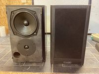 Picture of two bookshelf speakers, one with fabric grille removed showing mid/woofer and tweeter drivers and front bass port hole.