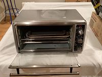 Front view of Hamilton Beach toaster oven showing stainless steel body, glass door open with two cooking trays inside, and control panel with mechanical knobs for settings.