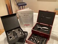 Two wine opener and aeration accessory sets in black and wood boxes with foam inserts. The New Sotheby's Wine Encyclopedia book standing upright next to them.