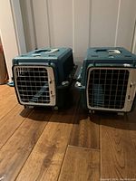 Two pet shuttle crates side by side on a wooden floor showing the front metal doors and plastic body.