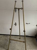 Full view of large antique brass freestanding easel standing on concrete floor, showing A-frame structure and crossbar.