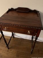 Photo of the antique wooden writing desk showing the carved top edge, brass drawer knobs, turned legs, and the raised backboard with decorative panel.