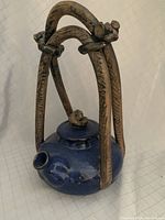 Blue glazed round pottery teapot with a four-legged distinctive curved ceramic handle structure with knot details at the top, on white fabric background