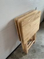 Three stacked folding pine TV tray tables against a wall, showing their thin profile and wood grain tops.