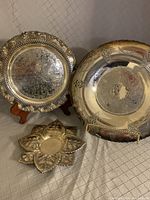 View of three silver trays displayed on checkered fabric. Left, leaf-shaped tray; center front, scalloped tray with grape design; right back, large round tray with engraved pattern and ornate edges.
