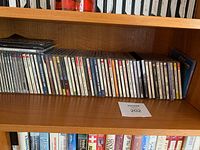 Wooden shelf with a single row of approximately 30 audio CDs mostly in jewel cases.