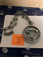 Full view of silver-colored metal necklace with a circular cougar head pendant on a thick chain, photographed on a white surface with a black and blue background.