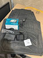 Two Garmin GPS devices, cables, mounting brackets and two large black rubber car mats shown on a ground surface.