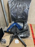 New, unassembled office chair parts wrapped in plastic on concrete floor near cardboard box containing small parts.