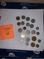 Overhead photo showing 17 assorted coins in various sizes and colors laid out in two horizontal rows on white paper with an orange MaxSold tag marked 34.