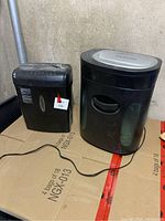 Two black shredders placed side-by-side on cardboard surface against a wall, showing full front and partial side views.