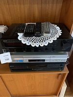 Stack of stereo components and remotes on a wooden shelf with white doily underneath.