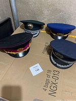 Four uniform hats placed on a table showing front and side details with badges and braiding.