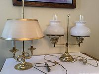 Two brass double lamps on a white surface; one lamp with candle style bulbs and fabric shade, the other with white gas burner style glass shades