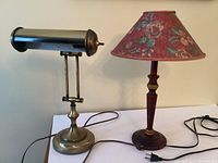 The two lamps side by side on a white surface showing the brass office lamp with cylindrical shade and the turned wood lamp with floral paper shade.