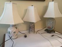 Three table lamps on a flat surface, showing the cream ceramic lamp with green floral transferware, stoneware lamp with glaze, and painted frosted glass lamp with brass base, all with white fabric shades.