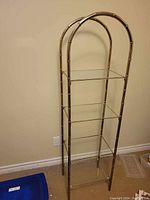 Full view of the brass and glass 4-shelf etagere against a wall, showing the bamboo-style segmented brass frame with two rounded arch tops and four glass shelves.