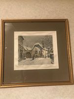 Framed painting on silk showing a winter village scene with snow-covered buildings and two figures walking down the street, matted and framed behind glass with a wooden gold-tone frame.