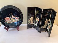 Front view of the painted wooden round plate on a stand and the adjacent four-panel lacquer folding screen with painted and raised figures of Japanese women.
