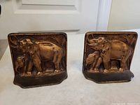 Front view of the pair of patinated metal elephant bookends showing adult elephant with calf in relief on each.