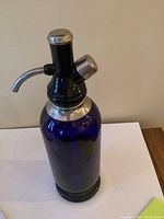 Cobalt blue glass soda siphon with metal top and spouts, standing on a flat surface