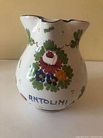 White ceramic floral jug with green, red, blue, and yellow painted flowers, labeled Antolini, showing detailed floral pattern and shape