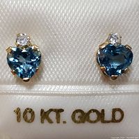 Close-up photo of the heart-shaped blue topaz earrings with a small clear gemstone, displayed on a white satin holder marked '10 KT. GOLD'
