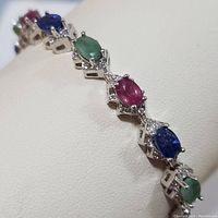 Close-up angled view of the bracelet showing alternating sapphire, emerald, and ruby stones in sterling silver setting with diamond accent stones.