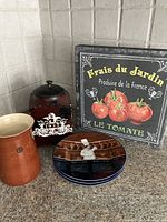 Photo of 3 salad plates illustrated with chefs in a kitchen by Guy Buffet, a black and red retro coffee tin, a brown pottery container, and a tomato-themed sign with French text.