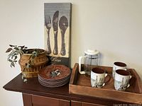 Photo showing the French press, three Eiffel Tower mugs, six dessert plates with Guy Buffet artwork, woven basket with plant, and kitchen sign with silverware design.