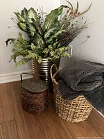 Photo of large metal container with faux greenery, small brown woven metal container beside a larger light brown woven basket with a grey throw blanket inside.