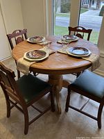 Full round dining table with leaf installed surrounded by 4 wooden chairs, each with black leatherette seats. Four place settings arranged on the table with decorative plates, white octagonal plates underneath, and glassware.