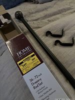 Photo of the boxed drapery rod with two matching oil rubbed bronze mounting brackets on a fabric surface. Box label indicates brand Home Decorators Collection and features a yellow sticker with pricing and rod size 36-72 inch.