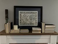 Full mantel display showing framed old-style map, decorative books stacked and lined, candleholder with candle on left side