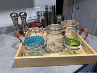 Six assorted glass storage jars with metal hinged lids on a wooden tray with handles shown on kitchen counter alongside salt and pepper shakers.