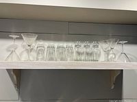 Wide shelf view showing clear martini glasses, short tumblers, Beck's beer glasses with logo, and several stemmed wine glasses arranged in a row