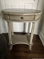 Front view of a decorative hall table with an oval top, turned legs, a single drawer with metal knob, and bottom shelf.