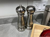 Two stainless steel salt and pepper grinders with rounded tops and curvy design placed on kitchen countertop near electrical outlets.