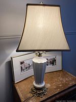 Tall ceramic table lamp with a cream-colored bell-shaped lampshade, sitting on a wooden surface with framed artwork behind.