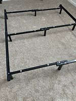 Black adjustable metal bed frame assembled on carpeted floor, showing framework, multiple supporting legs, and adjustable bars.