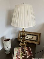 Photo showing brass-colored lamp with white pleated lampshade, textured vase, and framed 'Relax' sign on dark tabletop.