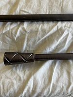 Photo of a dark-colored metal drapery rod segment with decorative crisscross pattern finial on one end, showing the rod's brushed finish.