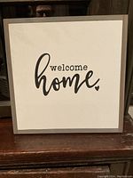Front of the sign showing 'welcome home' painted in black on a white background with grey wood frame.