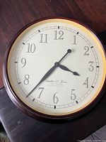 Top-down view of the round wood-framed wall clock, showing the white face with black numerals and clock hands along with brand text.