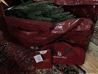 Artificial Christmas tree packed inside a red carry bag with Balsam Hill branding and accompanying small carry bag for tree stand. Catalog card beside items.